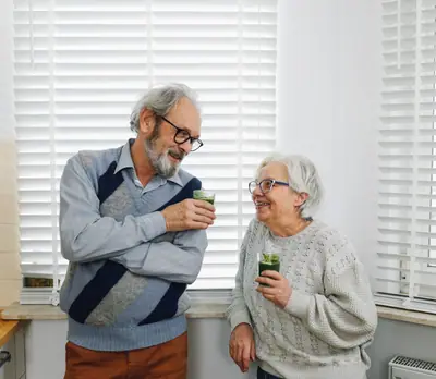 Casal de idosos ri na cozinha segurando copos de suco verde