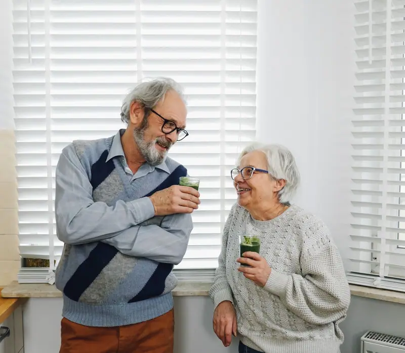 Casal de idosos ri na cozinha segurando copos de suco verde