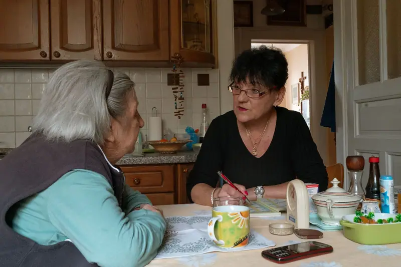 Mulher idosa e cuidadora conversam à mesa da cozinha, com chá e remédios organizados